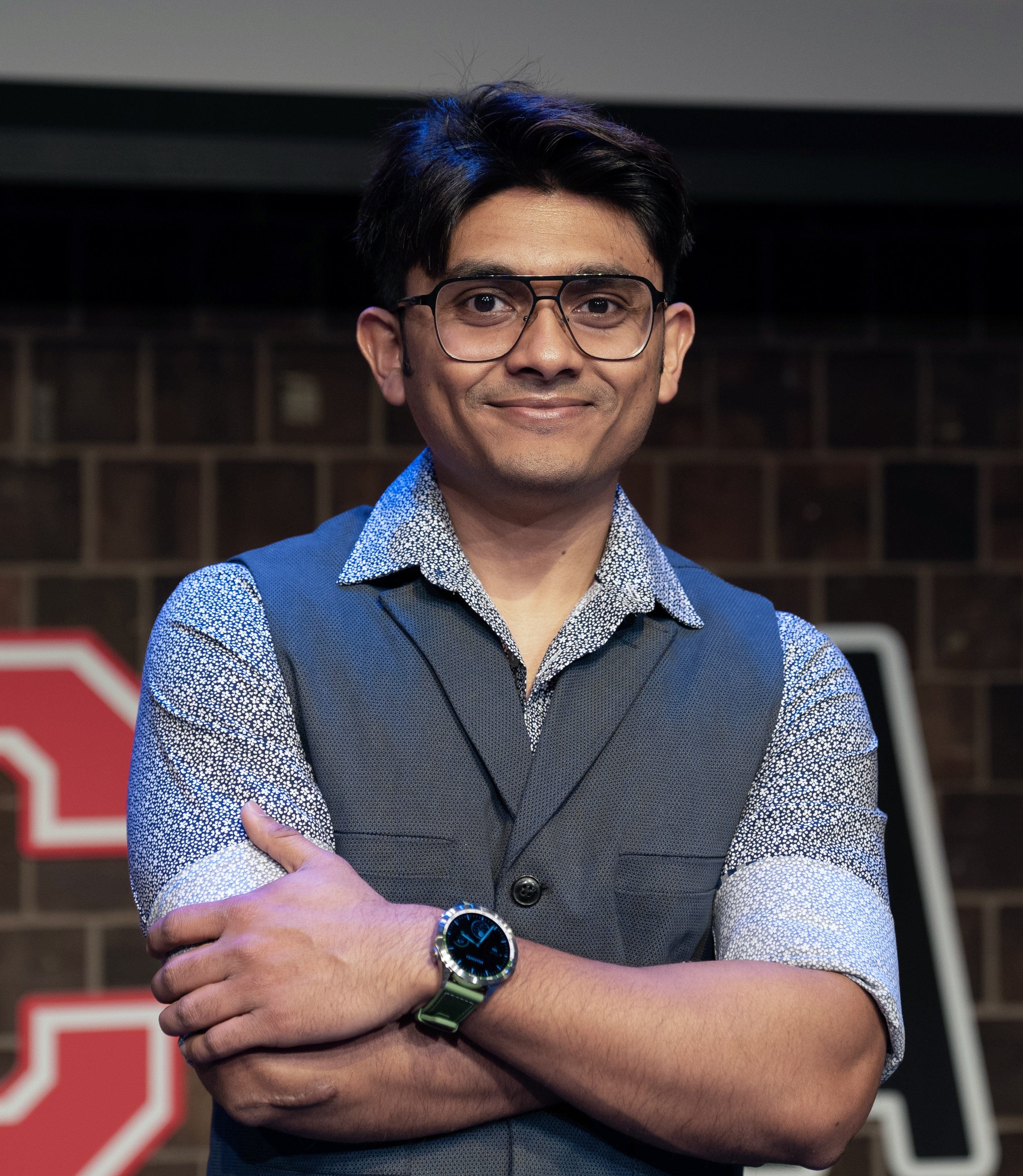 A man with a white shirt, blue vest, and a green watch. Crossed arms and smiling to the camera.